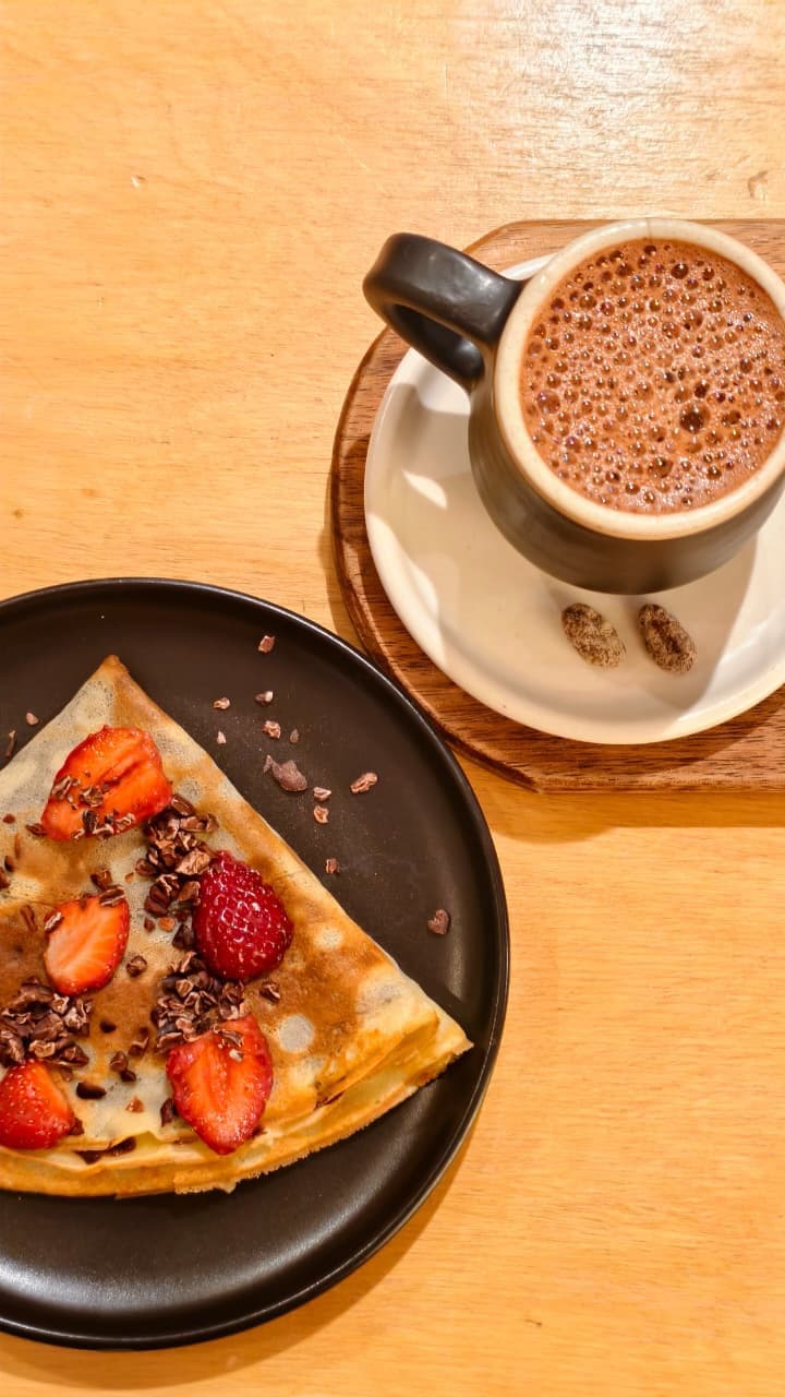 Una taza de chocolate caliente sobre una mesa de madera junto a un plato con crepa cubierta de fresas, frambuesas y nibs de cacao sobre un plato oscuro