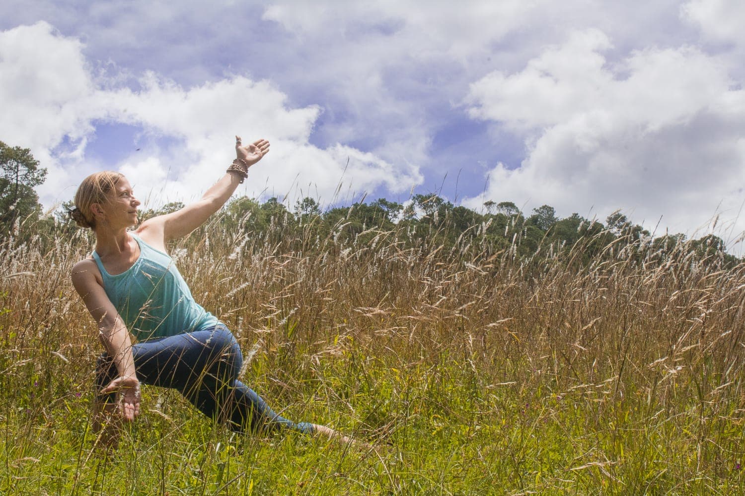 A woman in a turquoise sleeveless top and blue jeans performs a graceful yoga pose in a tall grassy field under a blue sky with scattered clouds; one arm raised and the other extended toward the ground, with trees in the distant background.