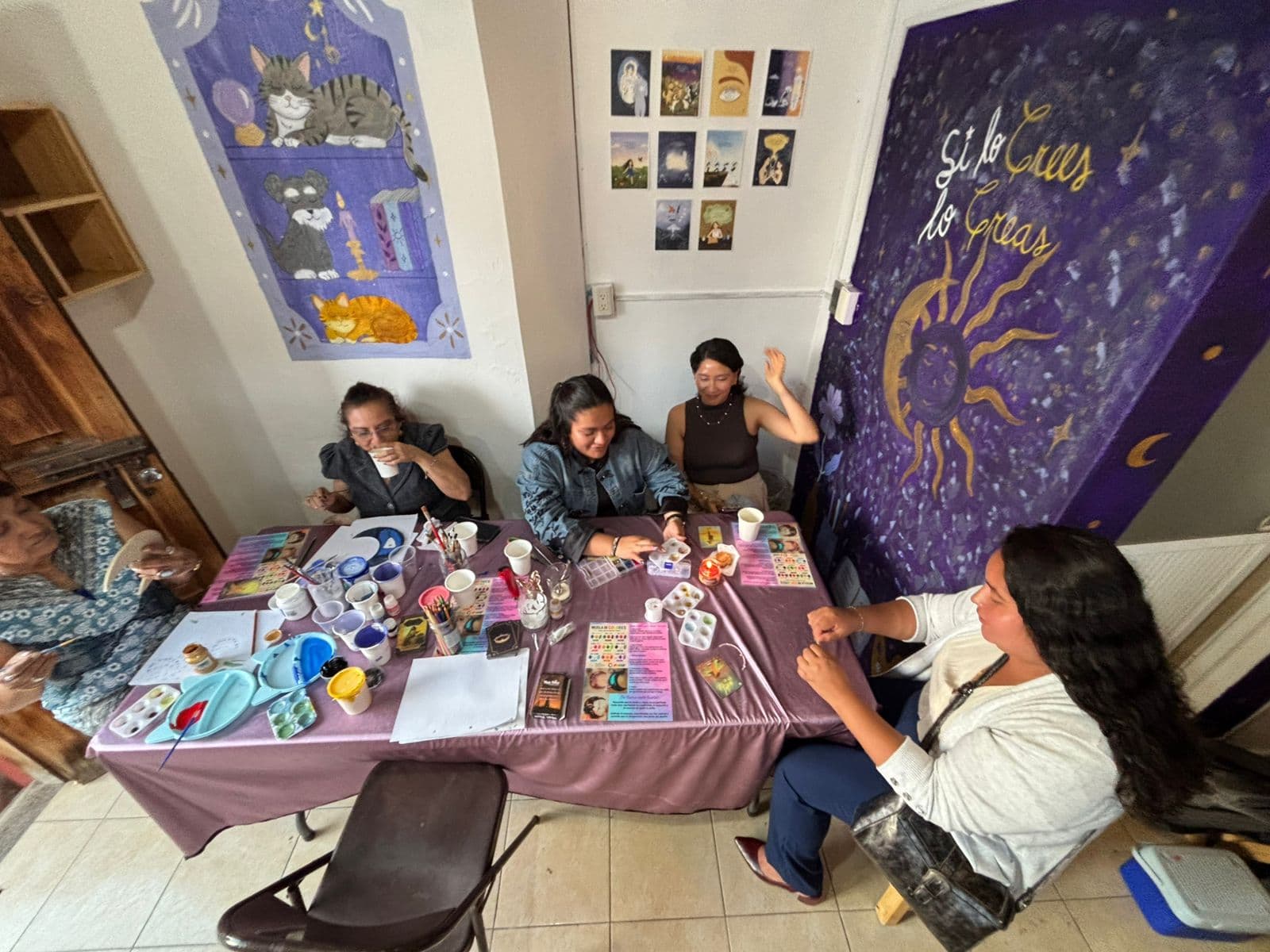 A group of five people sit around a rectangular table covered with a pink tablecloth engaged in arts and crafts. Supplies such as cups, brushes, palettes, papers, and small cards are spread across the table. The wall behind features a tall