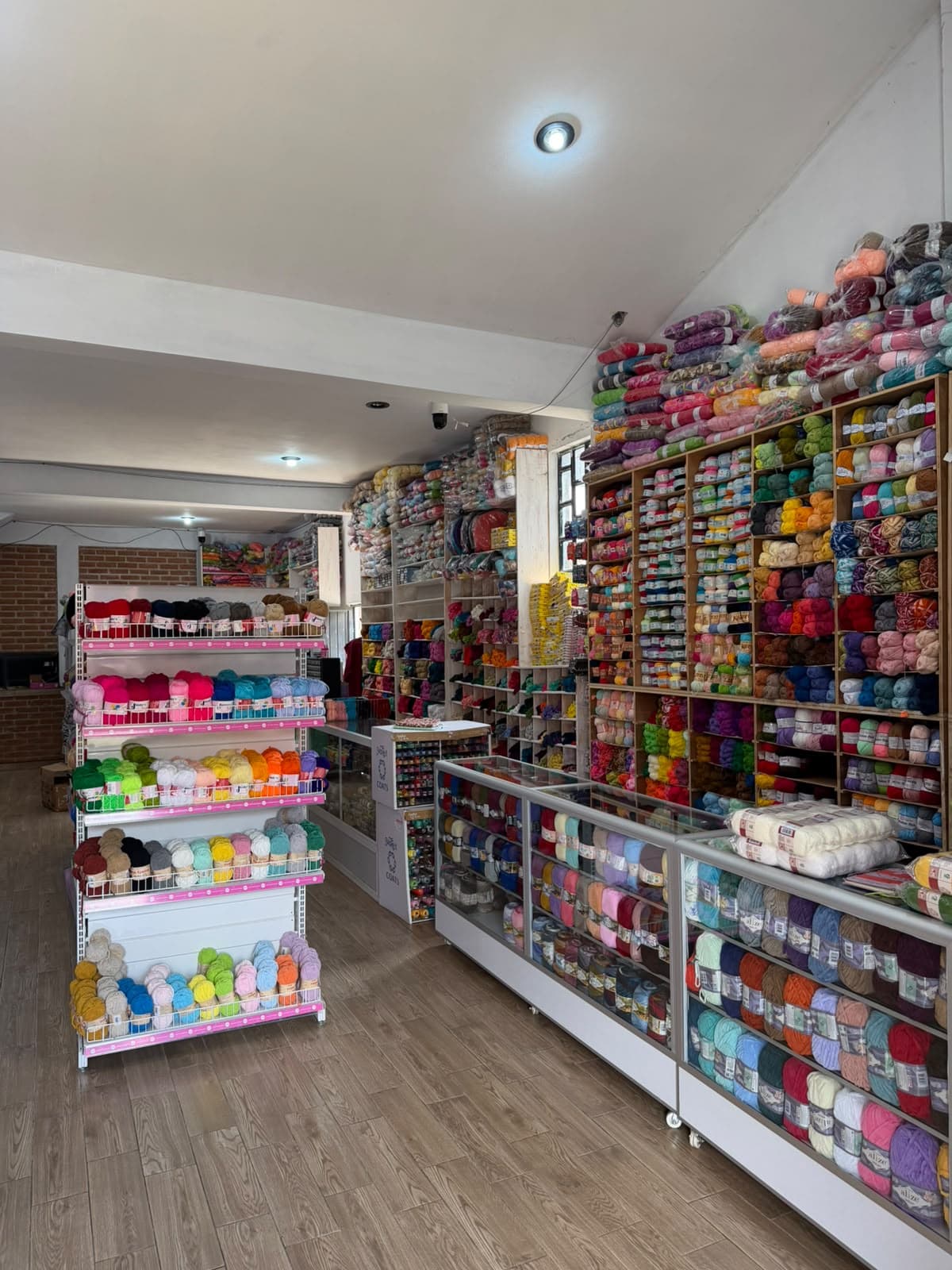 Interior of a yarn shop with tall shelves filled with colorful skeins and balls of yarn. A central display rack holds pastel-colored yarns, and long glass-fronted cabinets along the right side store more skeins. Wooden floor, white walls, a