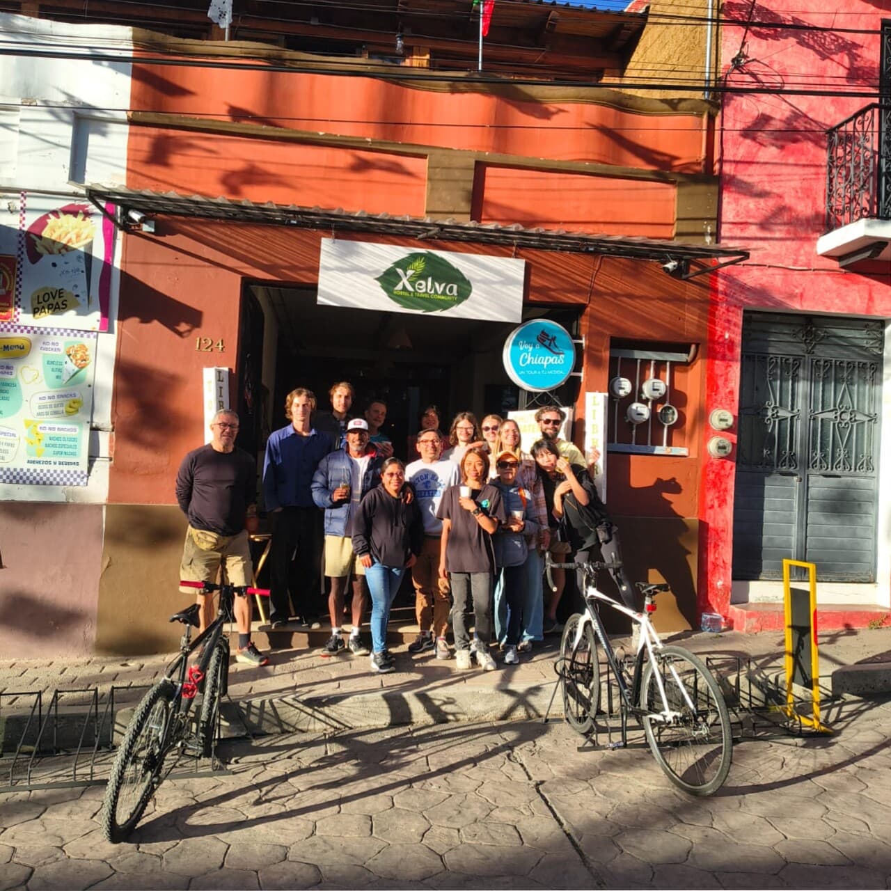 Group of people standing in front of a storefront with a sign that reads Xelva Hostel & Travel Community. A blue circular sign for Chiapas is visible. Bicycles are parked outside and there are colorful buildings with orange and red walls.