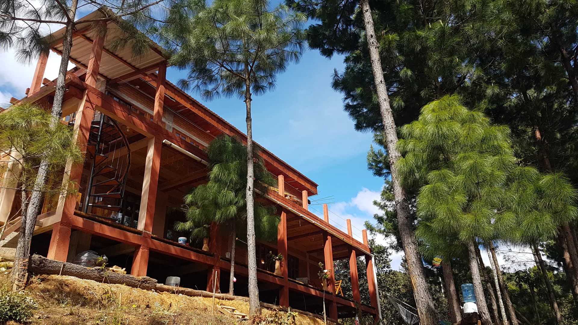A two-story wooden building under construction or renovation nestled among tall evergreen trees. The structure has exposed red-brown framing, open balconies, and a spiral staircase visible on the left side. Pine trees surround the site; a h